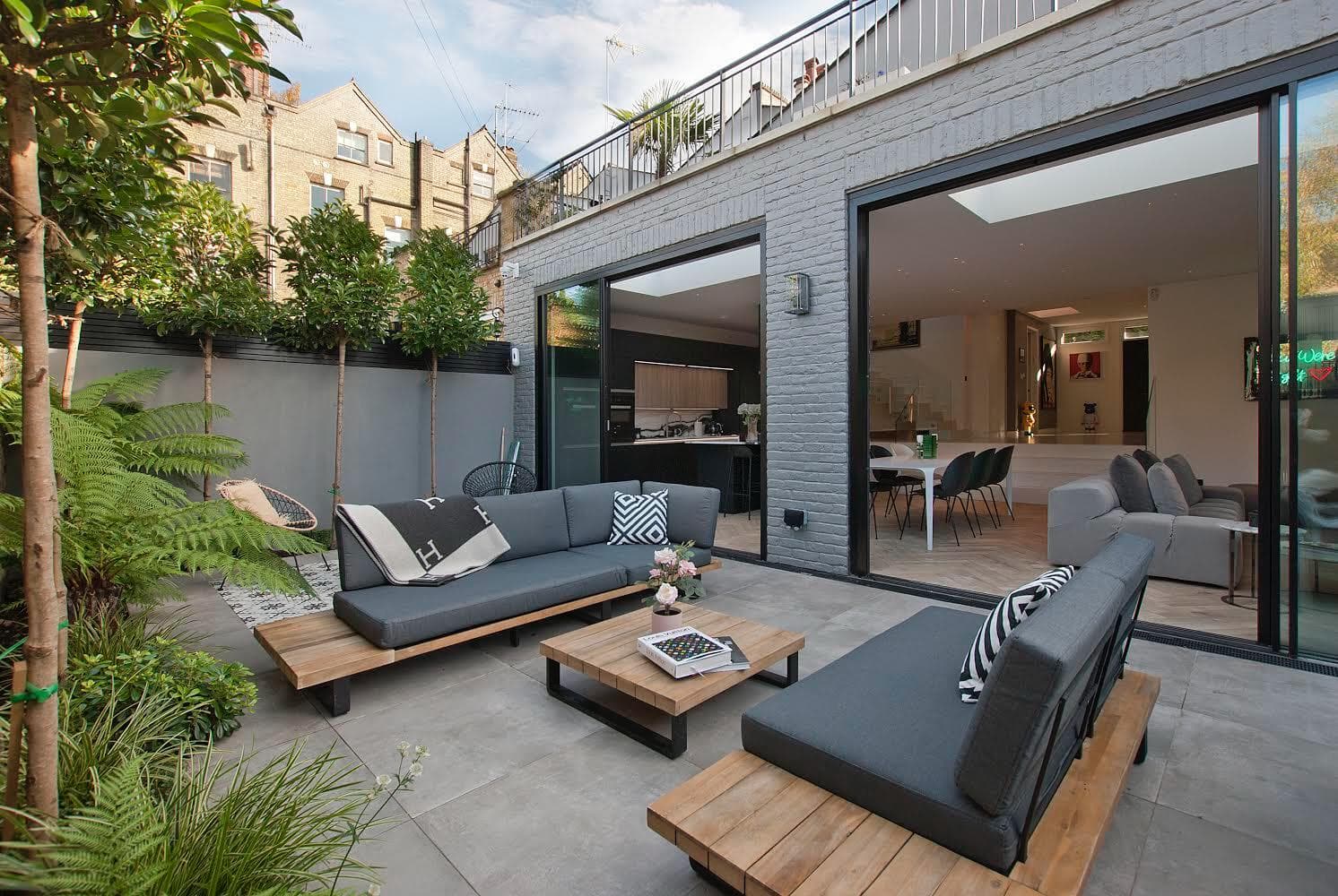 Modern patio with grey sofas, wooden furniture, and lush greenery outside sliding glass doors.