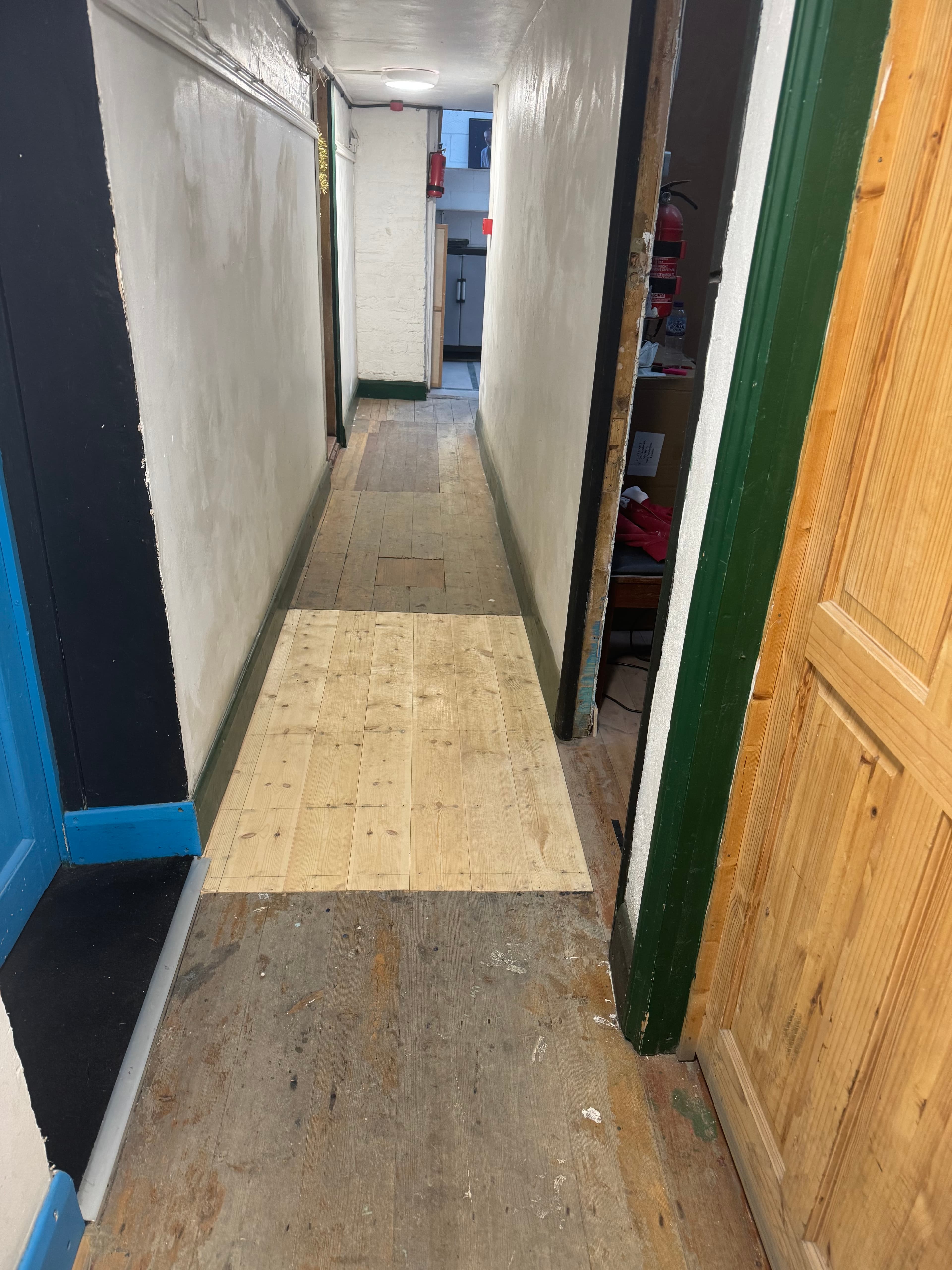 Narrow hallway with three distinct sections of wooden flooring, white walls, and colorful doorframes.